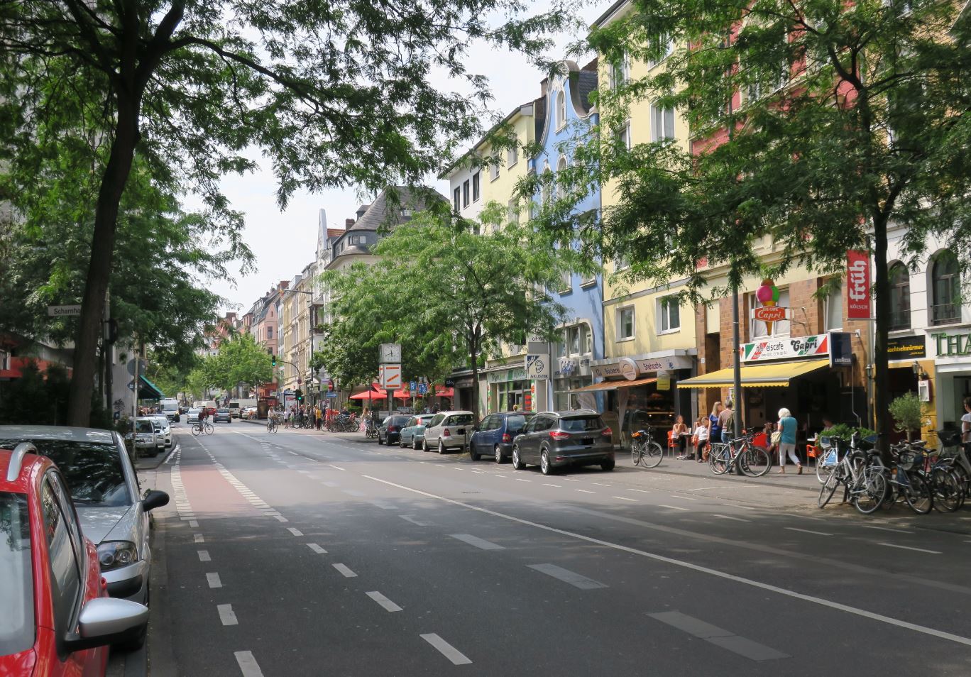 Auf dem Bild ist die Neusser Straße zu sehen. Es gibt parkende Autos und Menschen, die zu Fuß gehen oder mit dem Fahrrad fahren.