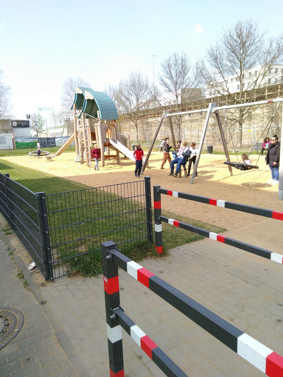 Spielplatz mit Schaukeln und einem großen Spiel- und Klettergerät, inklusive Rutsche. Auf dem Spielplatz sind Erwachsene und Kinder.