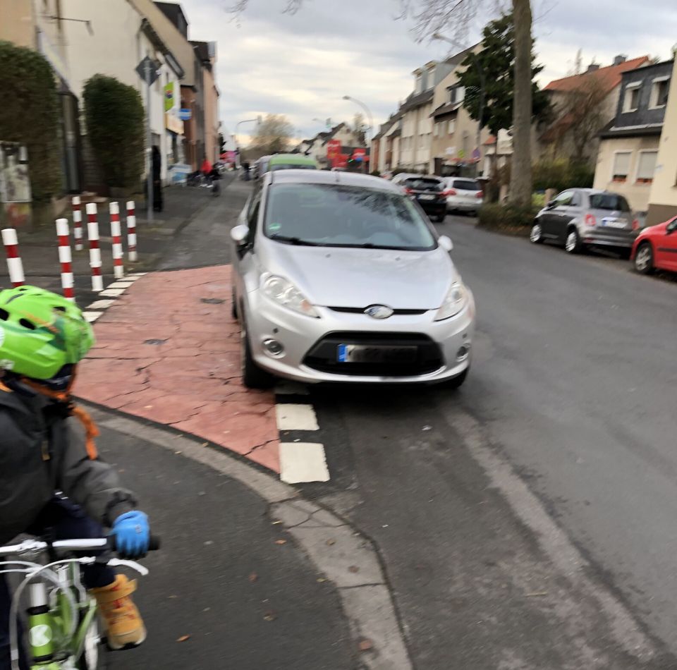 Links ist ein Kind auf dem Fahrrad. Ein Auto parkt auf dem Fahrradstreifen. 