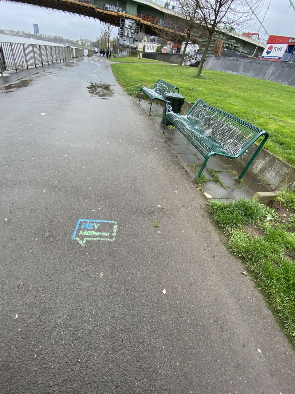Fußweg am Rhein. Rechts stehen zwei grüne Bänke, die mit Graffitis besprüht sind. Auf dem Gehweg ist in blauer und grüner Farbe das Logo "Hey Mülheim" gesprüht. 
