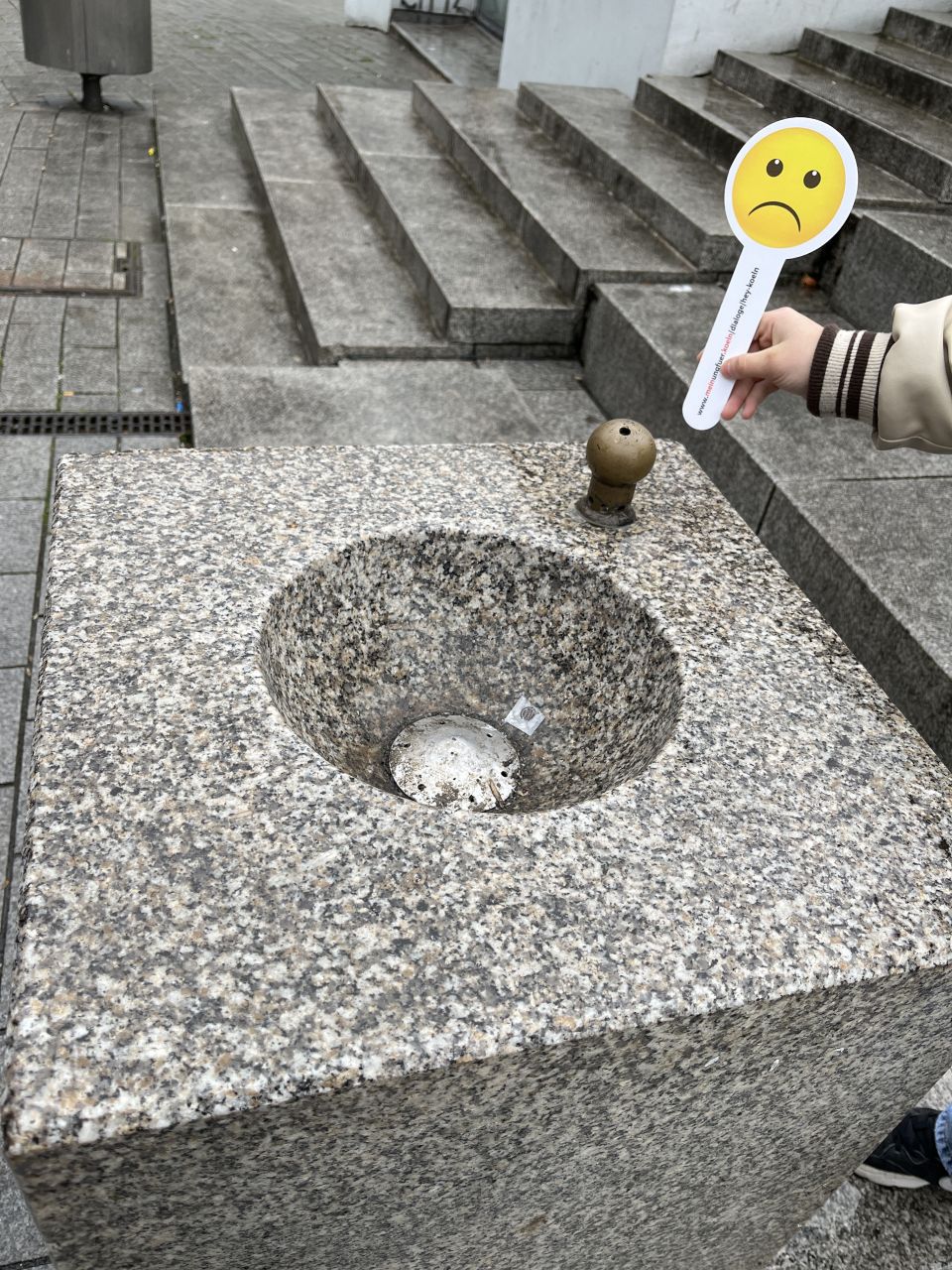 Ein Steinbrunnen, der nicht läuft. Ein Mensch hält eine Kelle mit einem traurigen Emoji ins Bild.
