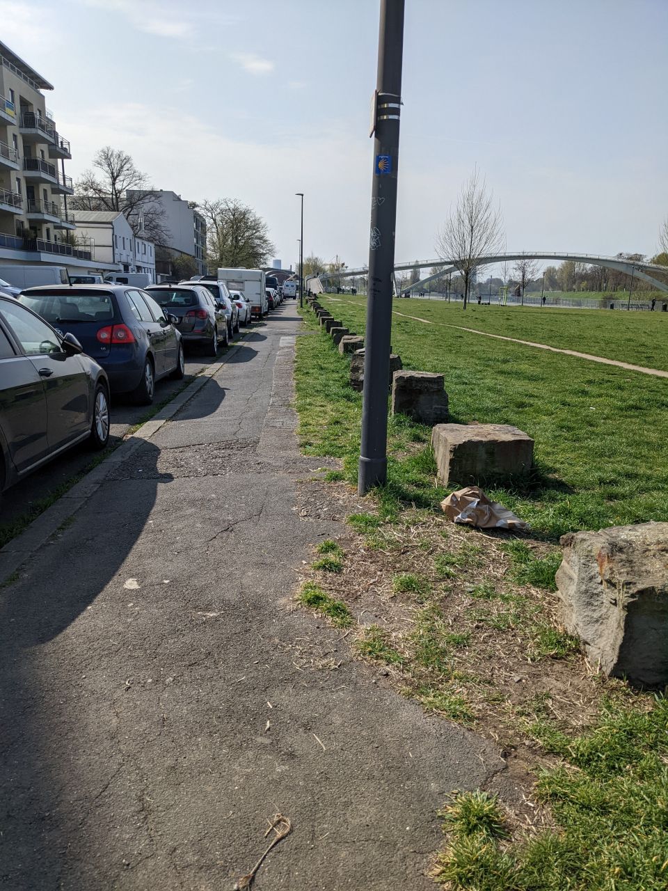 Gehweg am Rheinufer. Links vom Gehweg parken Autos. Rechts vom Gehweg ist eine grüne Wiese, die von großen Steinen abgegrenzt wird. 