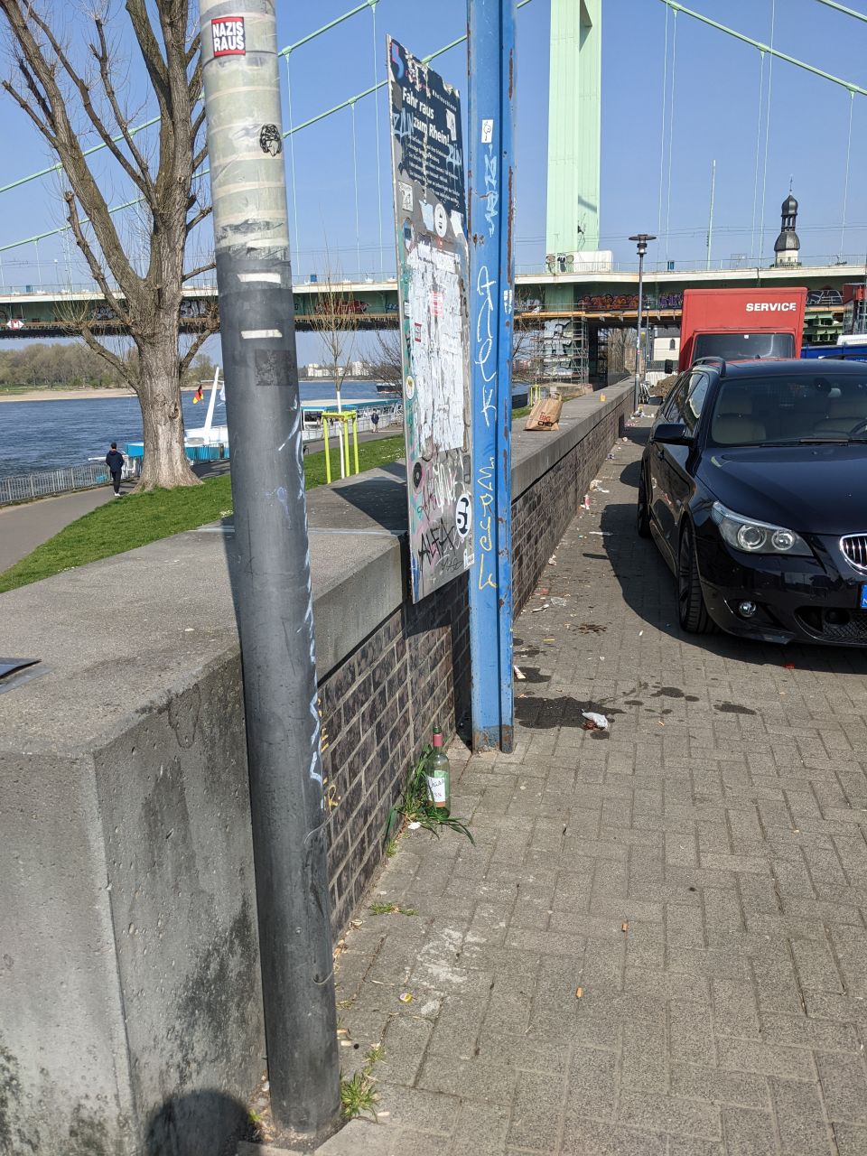 Weg am Rhein. Es parken Autos und links ist eine kleine Mauer. Im Hintergrund sieht man die Mülheimer Brücke.