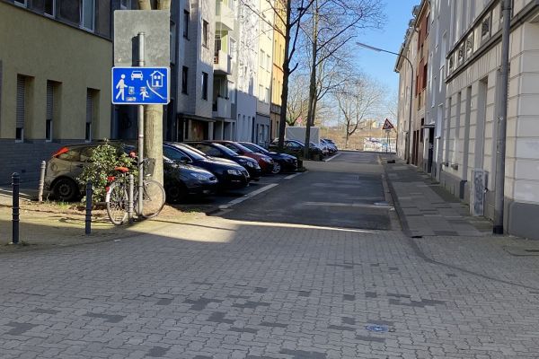 Zu sehen ist die Thumbstraße zwischen Kasernen- und Dieselstraße. Links parken Autos in Parktaschen. Rechts ist ein Bürgersteig. Ein Verkehrsschild zeigt an, dass dieser Bereich eine Spielstraße ist.