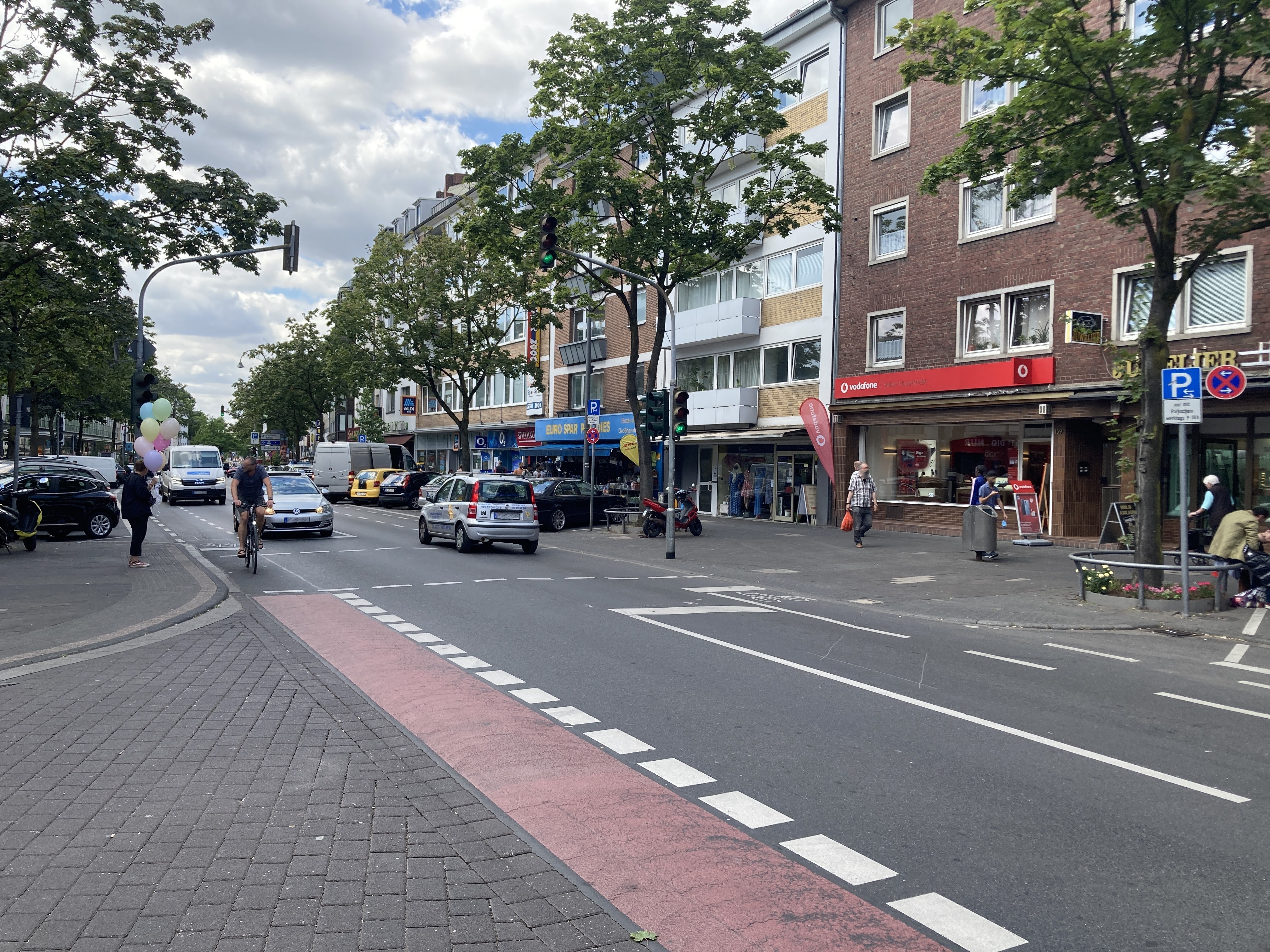 Ein Foto der Kalker Hauptstraße. Auf dem Gehweg laufen Fußgänger, auf der Straße fahren Autos. Eine Frau mit Luftballons wartet an einer Ampel. Auf deem Fahrradweg fährt ein Fahrradfahrer. Auf der Straße gibt es einige Ladenlokale und auf den Parkplätzen stehen Autos. Zwei Motorroller parken auf dem Gehweg. Der Gehweg ist zudem von Bäumen unterbrochen.