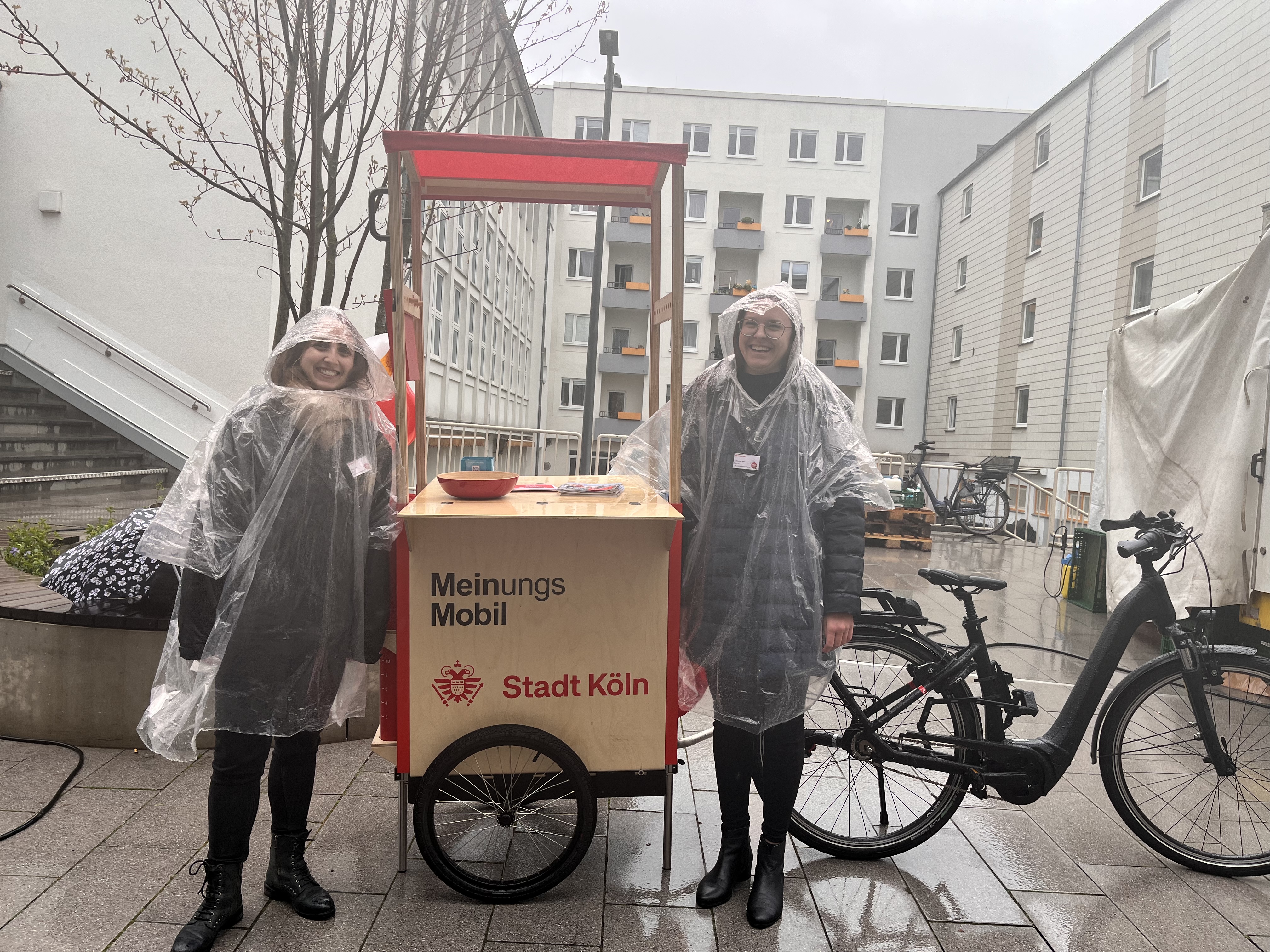 Zwei Mitarbeitende des Büros für Öffentlichkeitsbeteiligung stehen neben dem MeinungsMobil. Es regnet.