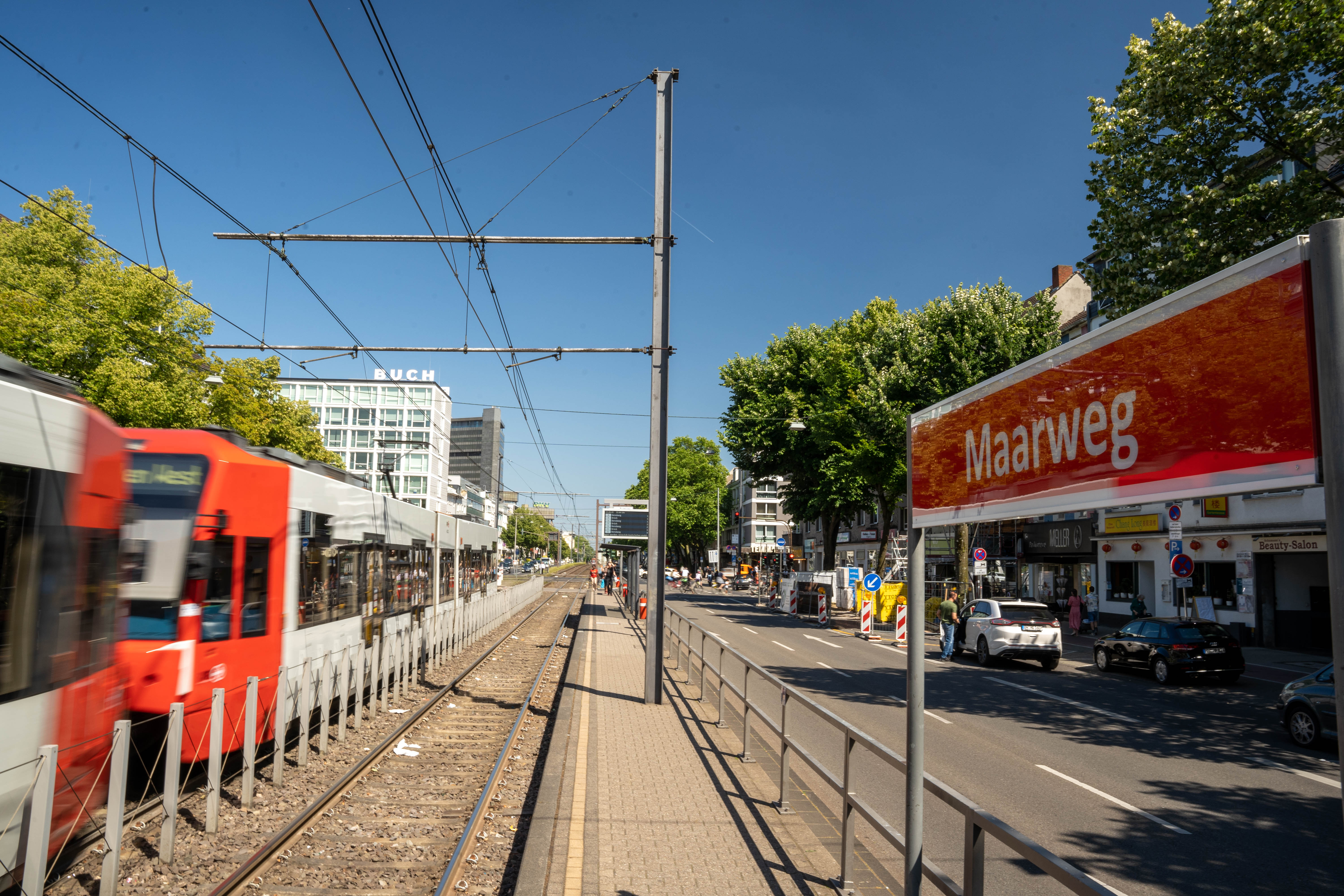 Seitlicher Blick vom Bahnsteig auf das Haltestellenschild Maarweg mit Straßenbahn im Hintergrund