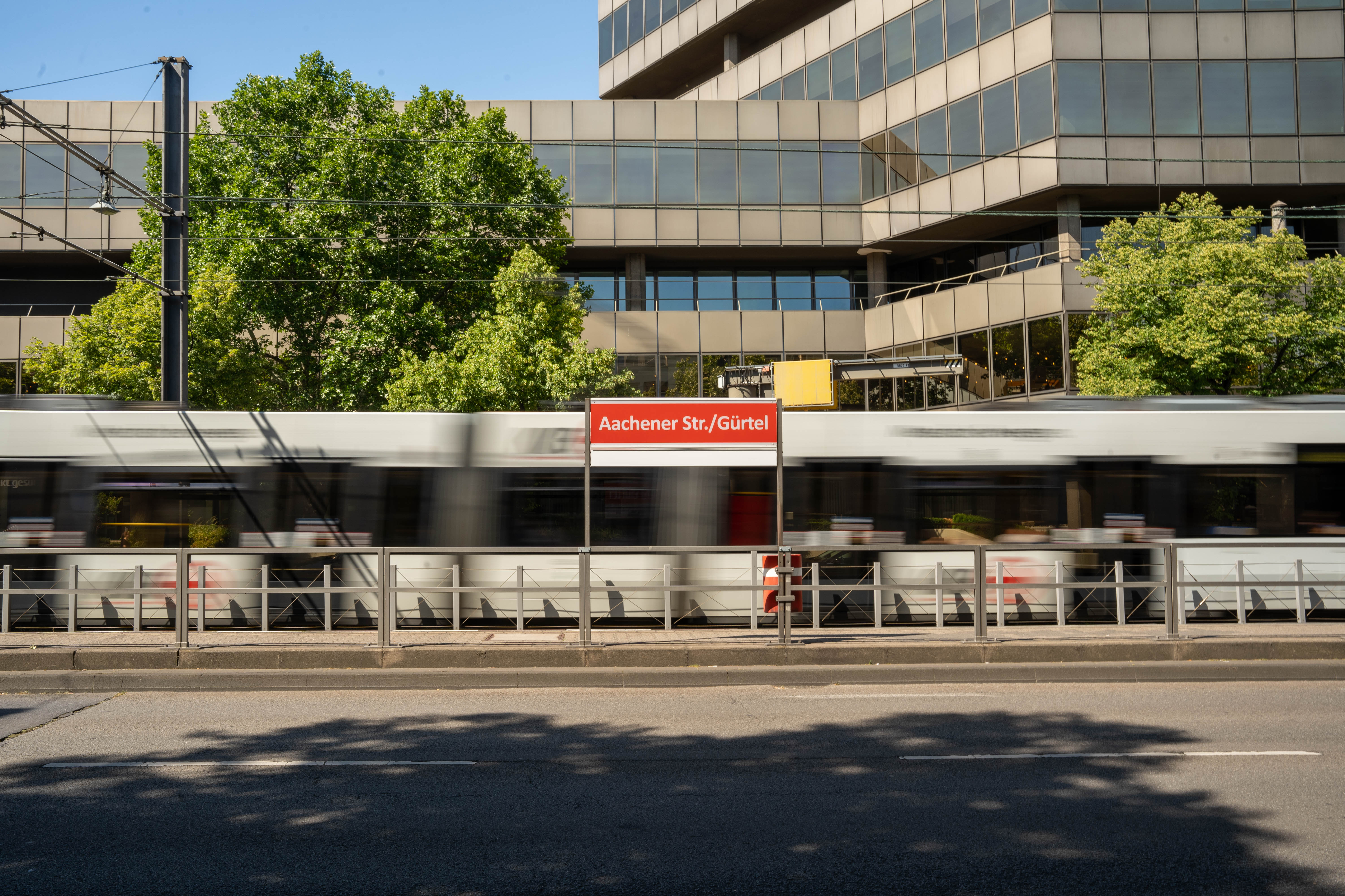 Blick von der gegenüberliegenden Straßenseite auf das Haltestellenschild Aachener Straße/Gürtel mit fahrender Straßenbahn im Hintergrund