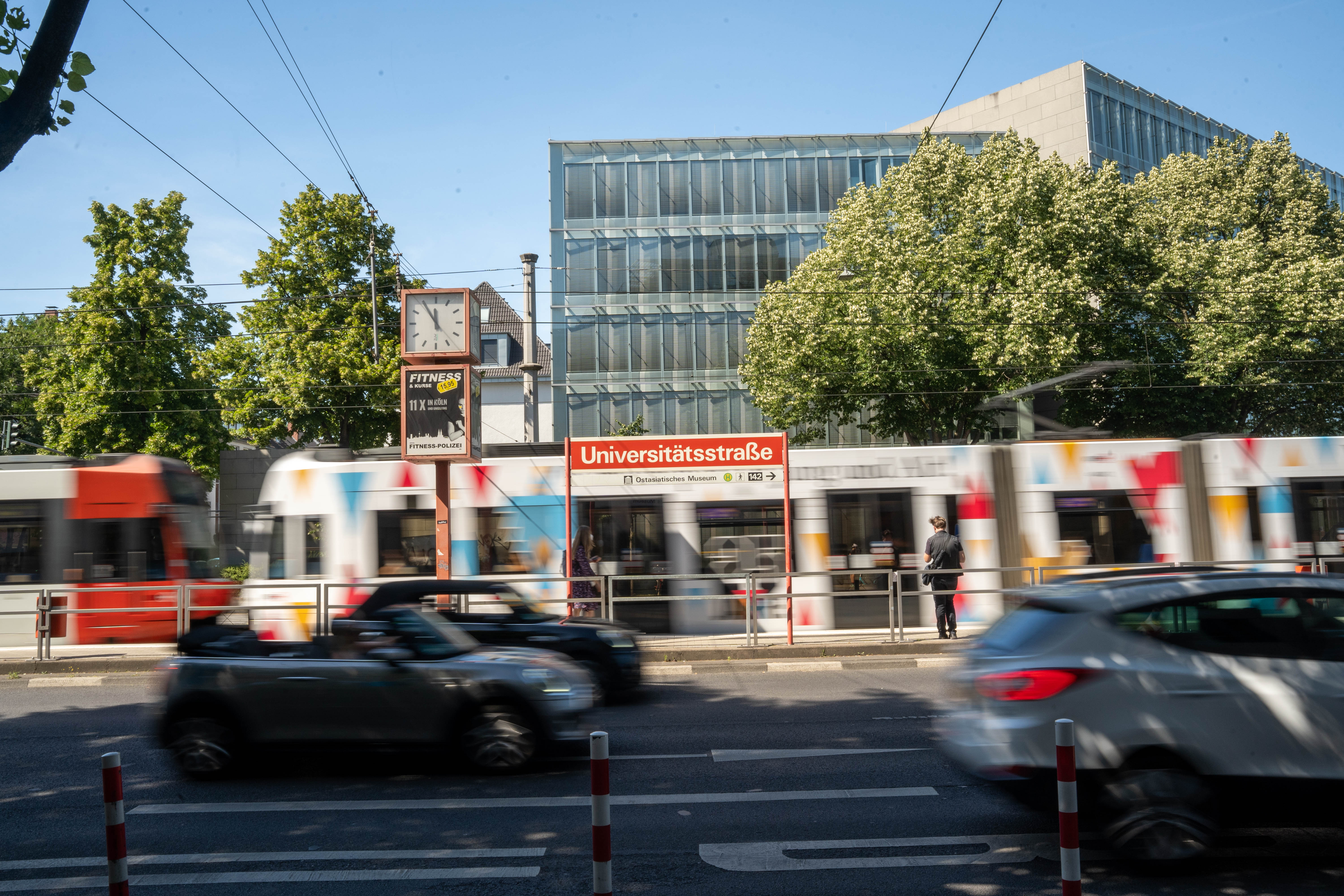 Blick von gegenüberliegender Straßenseite auf das Haltestellenschild Universitätsstraße mit Verkehr im Vordergrund und einer Straßenbahn im Hintergrund