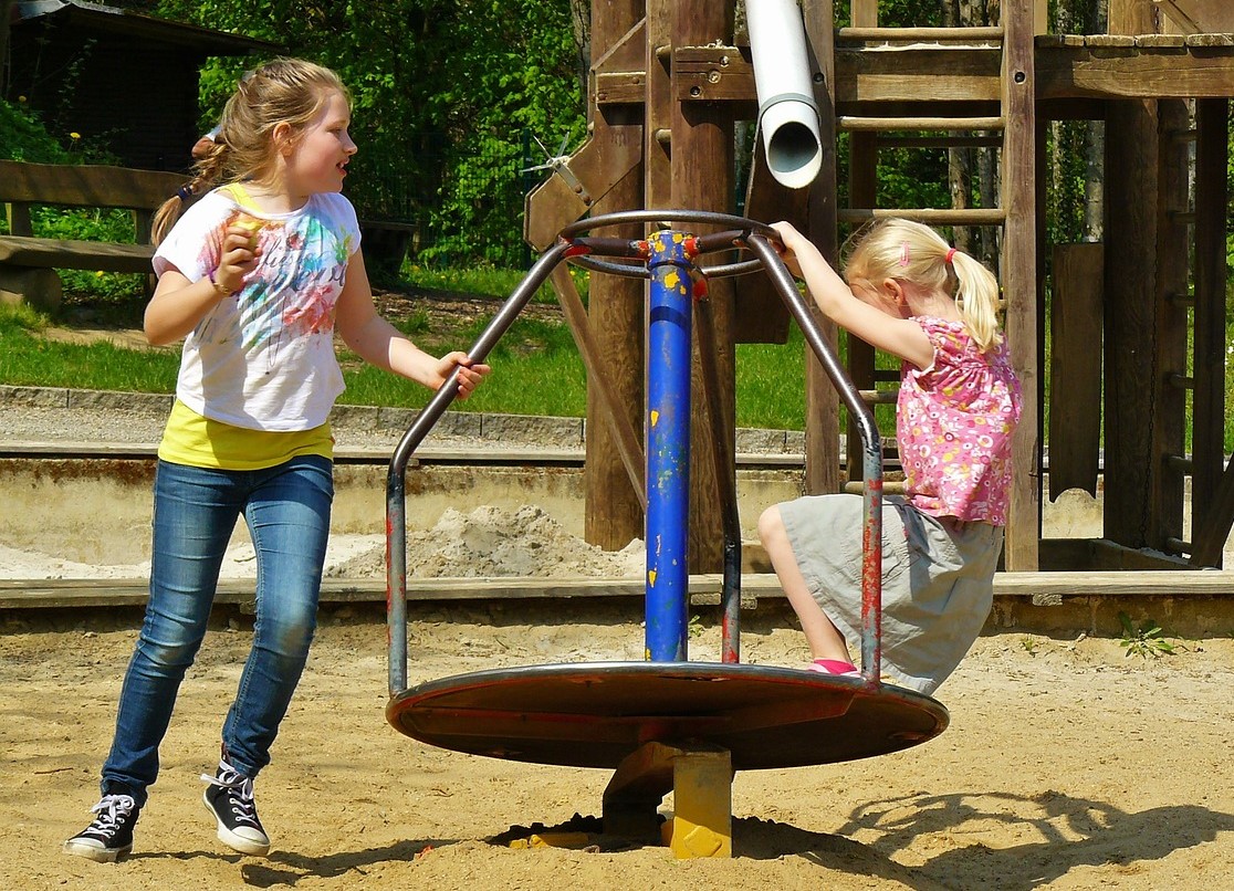 Zwei Kinder, die auf einer Drehscheibe spielen. Sie halten sich am auf der Drehscheibe besfestigten Gestänge fest.