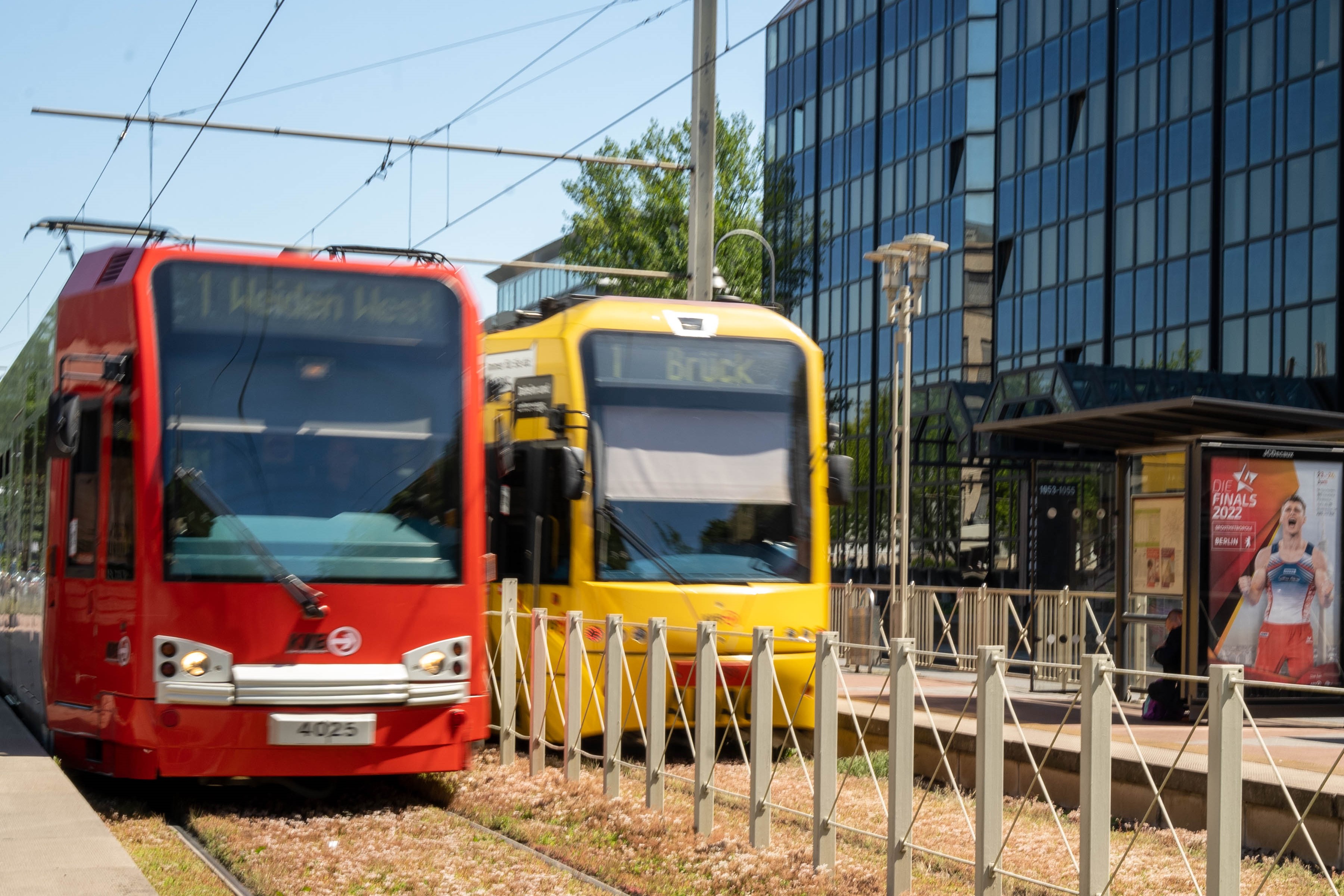 Blick auf zwei nebeneinanderstehende Bahnen der Linie 1 an der Haltestelle Mohnweg. Sie fahren in die entgegengesetzten Richtungen Weiden West und Brück. Zwischen den Bahnen verläuft ein Zaun, der die Gleise voneinander trennt.