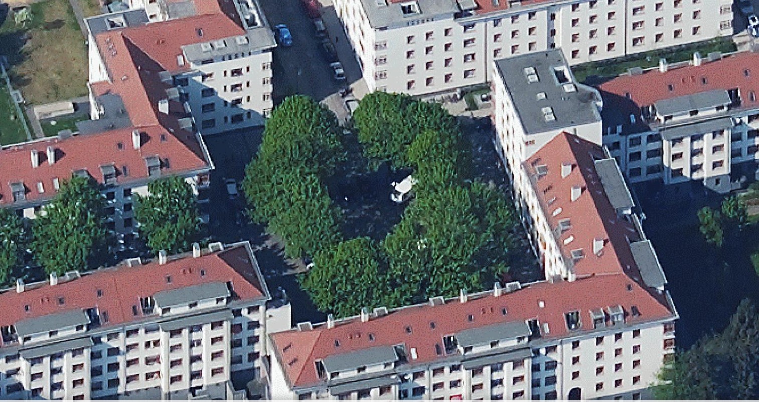 Naumannplatz in Köln-Riehl aus der Vogelperspektive. Der Platz ist von Bäumen umgeben. Um den Platz und die Bäume herum sind Mehrfamilienhäuser mit roten Dächern. Der Platz hat vier Zugänge über Straßen.