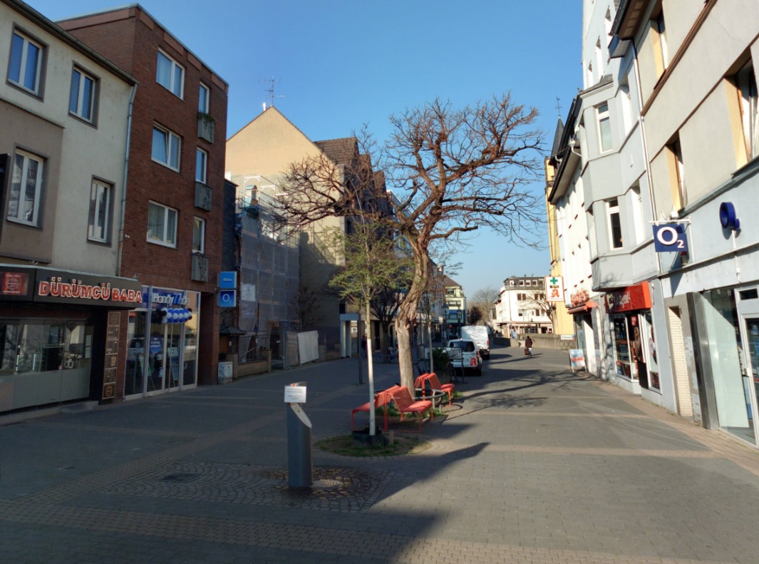 Ausschnitt der Bahnhofstraße auf welchem der Trinkbrunnen, Bänke sowie umliegende Gebäude mit Geschäften zu erkennen sind.