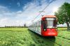 Eine rot-weiße Bahn der Kölner Verkehrsbetriebe fährt aus der Stadt Köln raus ins Grüne und die Natur. Im Hintergrund ist die Skyline von Köln mit dem Kölner Dom. Um die Bahn und die Schienen herum ist eine grüne Wiese mit kleinen, gelben Blumen und einem großen Baum.